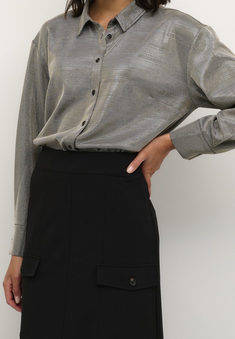 Femme portant une chemise argentée à manches longues boutonnée, rentrée dans une jupe noire taille haute avec de grandes poches boutonnées.