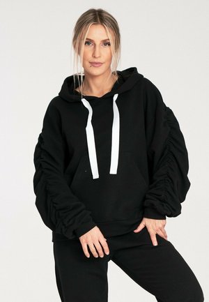 Czarna bluza z kapturem z przednią kieszenią, w luźnym fasonie, ze ściągaczami na rękawach i białymi sznurkami. Wykonana z miękkiego materiału, idealna na codzienne noszenie.