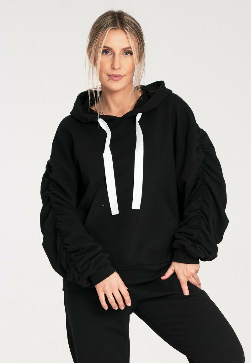 Czarna bluza z kapturem z przednią kieszenią, w luźnym fasonie, ze ściągaczami na rękawach i białymi sznurkami. Wykonana z miękkiego materiału, idealna na codzienne noszenie.
