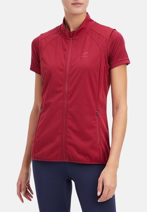 Woman wearing a sleeveless red zip-up vest over a short-sleeve shirt, paired with dark blue pants, against a plain background.