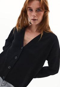 Mustaa neulottua kardigania, jossa on syvä V-pääntie, pitkät hihat ja viisi mustaa nappia edessä. Silkkinen rakenne ja rento istuvuus.