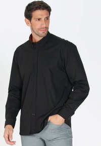 Chemise noire à manches longues avec col, en tissu lisse. Présente un détail de col intérieur rouge et une fermeture à bouton sur le devant. Forme ajustée.