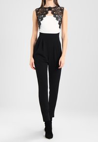 Combinaison noire avec un corsage blanc et un overlay en dentelle, design sans manches, taille haute et jambes fuselées. Texture lisse partout.