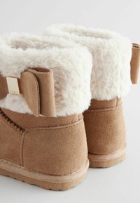 Brązowe zamszowe botki za kostkę z białym futrem sztucznym na górze i detalem w postaci kokardy. Posiadają przeszycia oraz teksturowaną gumową podeszwę zapewniającą przyczepność.