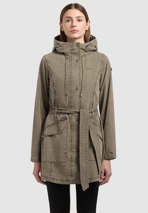 Mujer con parka verde oliva con capucha y cinturón, con bolsillos con solapa, de pie frente a un fondo claro y liso.