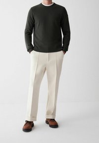 Tmavě zelený svetr s žebrovaným límcem, doplněný béžovými kalhotami s sklady a hnědými botami s černými podrážkami.