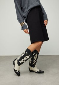Bottes de cowboy noires et ivoire avec des motifs complexes en forme de flammes, un bout pointu, un talon bloc bas et une texture en cuir lisse.