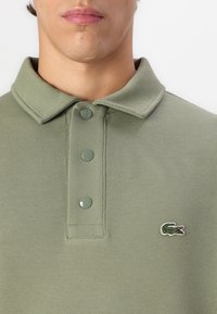 Camisa polo verde oliva hecha de tela texturizada, con cuello doblado y tres botones brillantes; presenta un pequeño logotipo de cocodrilo bordado.