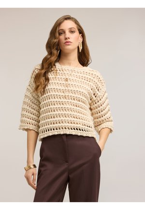 Frau mit welligem Haar, die einen beigefarbenen locker gestrickten Pullover, dunkle hoch taillierte Hosen, goldene Ohrringe, geschichtete Halsketten und Armbänder trägt.