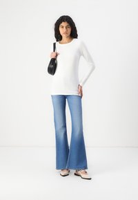 Top bianco a maniche lunghe, jeans svasati blu e ballerine beige. Tiene una borsa nera. Design semplice, silhouette aderente, texture del tessuto liscia.