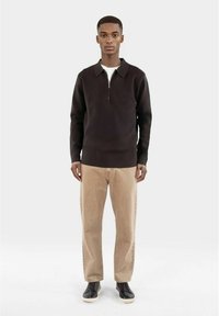 Svart hettegenser med glidelås og krage, beige rette bukser og svarte joggesko. Modellen står foran en nøytral bakgrunn.