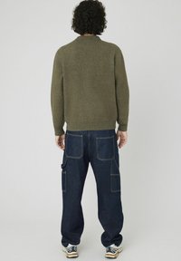 Pull en tricot olive avec un ourlet côtelé, associé à un pantalon cargo en denim bleu foncé avec plusieurs poches et des coutures visibles.