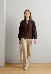 Brun langærmet bluse med bindebånd i halsen og samlet skuldre, parret med beige vidde bukser og en stor lysfarvet tote bag.