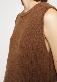Primer plano de una persona que lleva una blusa sin mangas de punto marrón texturizado con un escote redondo, sobre un fondo liso.