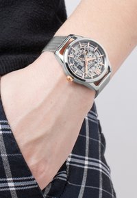 Skeleto laikrodis su sidabrine nerūdijančio plieno tinklelio juosta. Apvalus korpusas turi skaidrų ciferblatą, rodantį vidinius mechanizmus, rožinio aukso akcentais.