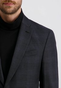 Mann trägt ein Blazer mit dunklem Karomuster über einem schwarzen Rollkragenpullover, zeigt Bart und Schnurrbart vor einem schlichten hellen Hintergrund.