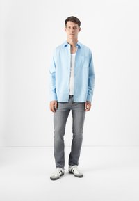 Ein junger Mann steht vor einem schlichten weißen Hintergrund und trägt ein hellblaues, kurzärmliges Hemd, ein weißes Tanktop, graue Jeans und Turnschuhe.