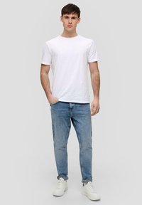 Biała koszulka krótki rękaw, jasnoniebieskie dżinsy, białe sneakersy. Model stoi prosto z rękami w kieszeniach, neutralne tło.