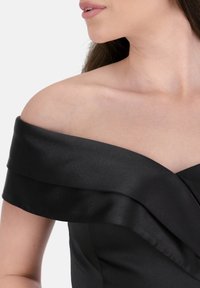 Svart satin klänning med off-shoulder-design, med lager av volanger och en slät textur som framhäver halsringningen och axlarna.