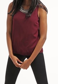 Top bordeaux senza maniche con un collo di perline testurizzato. Il tessuto è liscio e la vestibilità è rilassata, abbinato a pantaloni neri attillati.