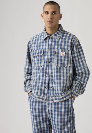 Vyras dėvi mėlynos ir baltos spalvos languotą mygtukais užsegamą švarkelį ir derančias kelnes, stovintis su rankomis kišenėse priešais paprastą foną.