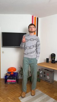 Pulôver cinza com um padrão de corrente, calças verde-claro e botas beige. O fundo apresenta uma televisão, uma lâmpada de mesa laranja e uma planta em vaso.