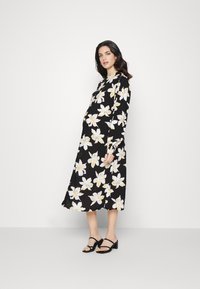 Vestido midi preto com padrão floral branco e amarelo, mangas longas e corpete ajustado, combinado com sandálias de salto pretas com tiras.