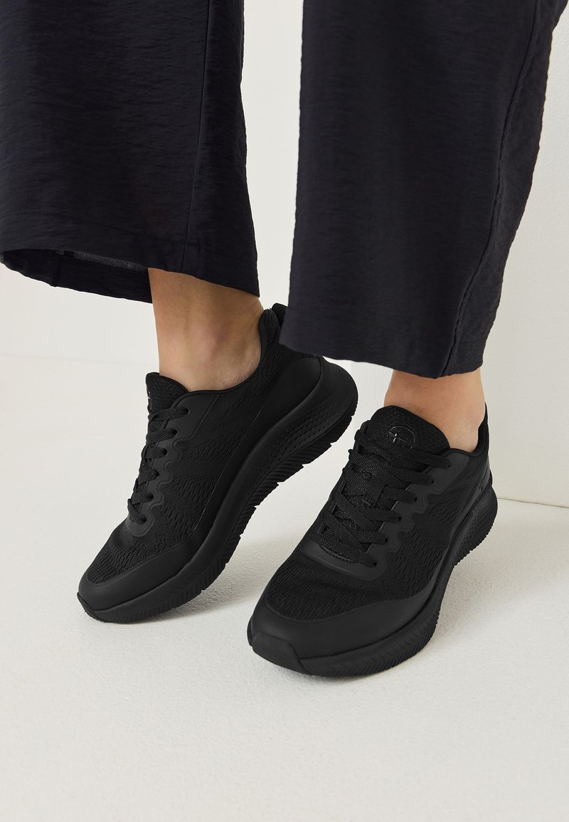 Personne portant des baskets noires avec des semelles texturées et un pantalon noir, debout sur une surface blanche.