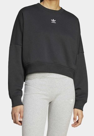 Nainen, jolla on musta lyhyt Adidas-collegepaita ja vaaleanharmaat trikoot, joissa on Adidas-logot, seisomassa yksinkertaisen taustan edessä.