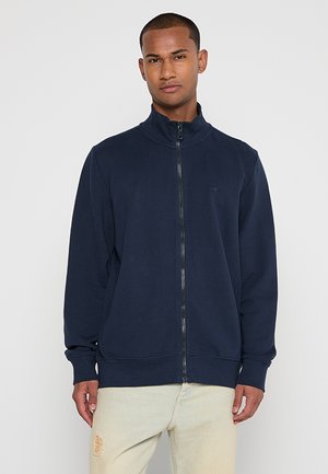 Námořnická mikina na zip vyrobená z měkké tkaniny, s vysokým límcem, bočními kapsami a hladkým povrchem. Jednoduchý design bez vzorů.