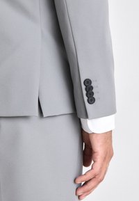 Traje gris a medida con una textura suave, que presenta cuatro botones negros en el puño y un puño de camisa blanca visible debajo.