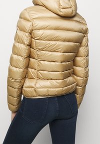 Gouden pufferjack met capuchon, voorzien van doorgestikte delen, glad satijnen oppervlak en elastische zoom. Gecombineerd met donkerblauwe spijkerbroek.