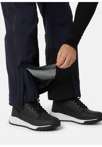 Marineblauwe waterproof broeken met ritssluitingen bij de enkels, gecombineerd met zwarte enkelboots met witte zolen en zwarte veters.