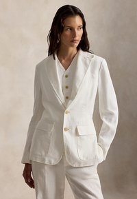 Γυναίκα με μακριά σκουρόχρωμα μαλλιά, ντυμένη με εκρού ραμμένο σακάκι, ταιριαστό γιλέκο με κουμπιά και παντελόνι, που στέκεται με το ένα χέρι στην τσέπη.