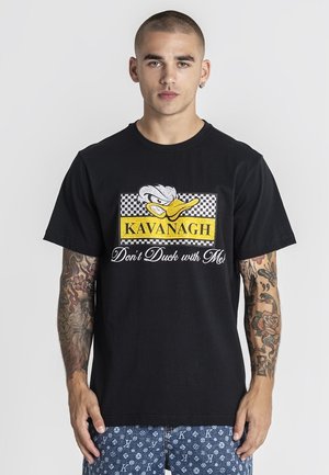 Giovane con capelli corti e tatuaggi su entrambe le braccia che indossa una maglietta nera con la scritta "Kavanagh Don't Duck with Me" e pantaloncini blu con motivi.