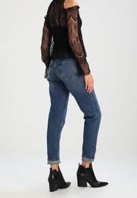 Top off-shoulder in pizzo nero abbinato a jeans in denim blu a vita alta. La modella indossa stivali neri alla caviglia. Dettaglio con maniche corte e risvolti.