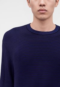 Jeune personne portant un pull en maille texturée bleu marine avec un col rond, présenté du cou jusqu'à la partie supérieure de la poitrine sur un fond uni.