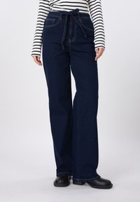 Σκούρα μπλε τζιν ψηλόμεσα με ζώνη από ύφασμα, φαρδιά πόδια και ορατές ραφές. Συνδυάζονται με μακρυμάνικο πουκάμισο ασπρόμαυρες ρίγες.