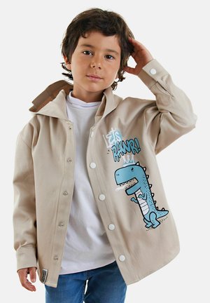 Beige button-up jack met een grote blauwe dinosaurusgrafiek, witte knopen en een zachte katoenstructuur, gelaagd over een wit shirt.