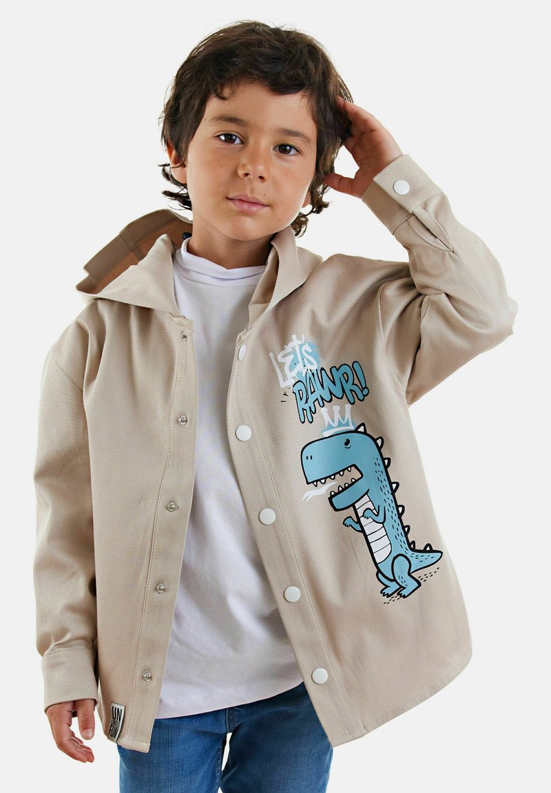 Giacca beige con bottoni, decorata con una grande grafica di un dinosauro blu, bottoni bianchi e una texture in cotone morbido, indossata sopra una camicia bianca.