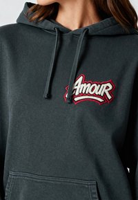 Felpa con cappuccio grigio scuro caratterizzata da un logo ricamato "Amour" in rosso e bianco. Presenta una tasca a marsupio e un cappuccio con lacci. Tessuto morbido e dal design testurizzato.