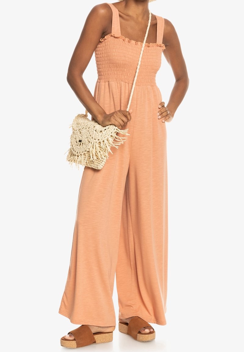 Jumpsuit v broskvové barvě se smockovaným trupem, širokými nohavicemi a volánkovými ramínky. V páru s tkanou kabelkou s třásněmi a hnědými platformovými sandály.
