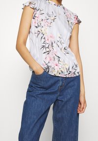 Blommig blus i rosa och svart blommönster på en ljusgrå randig bakgrund, med volangärmar och rund halsringning.