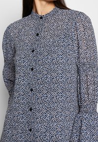 Femme portant un chemisier à manches longues boutonné avec un petit motif floral bleu, noir et blanc et un col montant.