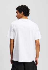 Weißes Baumwoll-T-Shirt mit kurzen Ärmeln, Rundhalsausschnitt und einem Etikett an dem Saum. Glatte Textur, lockere Passform und minimales Design.