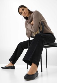 Bruine geribbelde trui, zwarte wijde jeans en zwarte flats. Model zit op een zwarte stoel met een geweven rugleuning in een ontspannen houding.