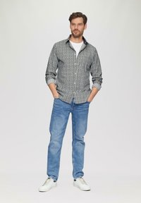 Hemd mit Knopfleiste in einem schwarz-weißen Muster, kombiniert mit hellblauen Jeans und weißen Sneakern. Das Modell steht mit den Händen in den Taschen.