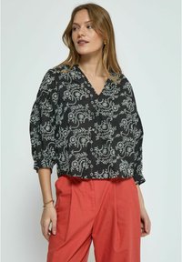 Schwarze Bluse mit weißen Blumenmustern, V-Ausschnitt und lockeren Ärmeln. Kombiniert mit korallfarbenen Hosen. Weicher Stoff, lässiges Design.