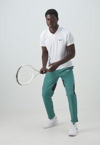Witte polo met groen Nike-logo, teal sportbroek met zwarte accenten, houdt een tennisracket vast, draagt witte sneakers.