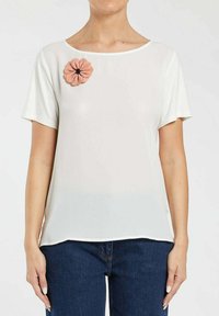 Hvid kortærmet top med rund halsudskæring, prydet med en tekstureret pink blomsterapplikation på venstre bryst. Blødt, let stof.